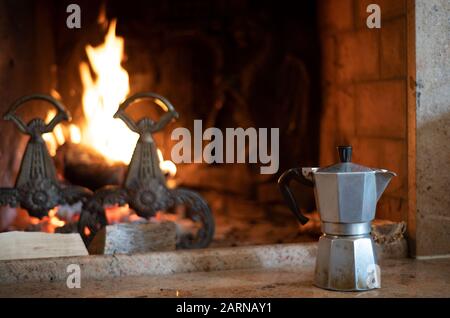 Italienische Kaffeekanne auf dem Herd in der Nähe eines brennenden Feuers mit alten Andironen in einem offenen Kamin in Nahsicht im Winter Stockfoto