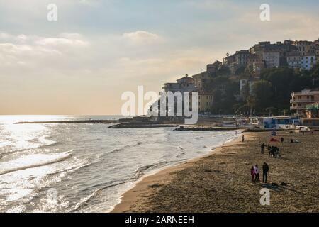 Der sandige Strand von Porto Maurizio mit den Menschen, die an einem warmen Wintertag die Sonne genießen, und die Altstadt von Parasio am kap, Imperia, Ligurien, Italien Stockfoto