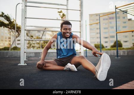 Portrait eines lächelnden Mannläufers, der auf dem Sportpark im Freien sitzt und Beine streckt, die sich auf das Lauftraining vorbereiten Stockfoto