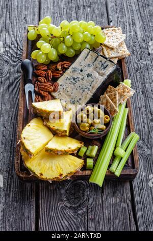 Blaukäse serviert mit Pekannuss, grünen Trauben, Selleriestangen, Keksknackern, Ananasscheiben und Oliven auf einem unschönen Holztablett auf einem rustikalen Tisch, ver Stockfoto