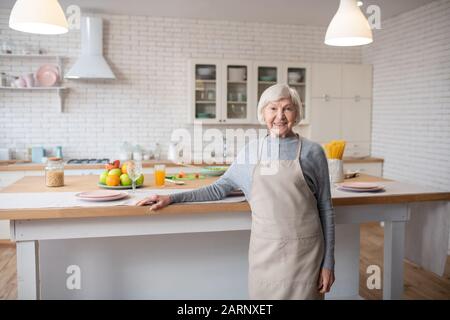 Weißhaarige Frau in Schürze, die vor dem Küchentisch steht. Stockfoto