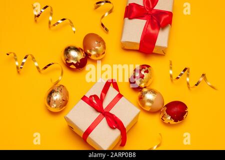Überraschungsgeschenkkasten in Handwerkspapier und mit rotem Bogen und goldenen Eiern auf gelbem Hintergrund. Glitzernder Serpentin. Osterfeier Stockfoto