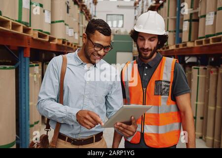 Junger Geschäftsmann, der Daten auf dem Touchpad für Lagerarbeiter mit Hardhat und orangefarbener Jacke anzeigt Stockfoto