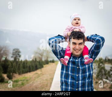 Ein kleines Mädchen, das an einem übergiebelten Tag auf den Schultern ihres Vaters in einem Park reitet. Stockfoto