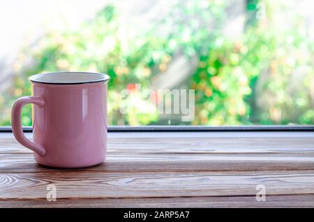 Eine pinkfarbene Tasse Tee steht auf einem Holzfensterschweller vor dem Hintergrund eines Sommergartens Stockfoto