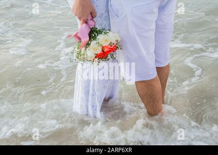 Der Blick auf die frisch vermählten Beine am Strand. Der Blick auf den perfekten Hochzeitsstrauß. Hochzeit Blumen Blumenstrauß in der Hand der Braut. Bräutigam. Stockfoto