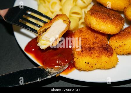 Ein Stück Nuggets auf einer Gabel und ein weißer Teller mit gebratenen Hühnerhuhn Nuggets, Pasta und Ketchup auf dunklem Hintergrund. Nahaufnahme Stockfoto