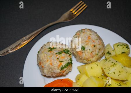 Zwei Fleischfleischbällchen mit gekochten Kartoffeln und gekochten Karotten auf einem weißen Teller mit einer Gabel auf dunklem Grund. Nahaufnahme Stockfoto