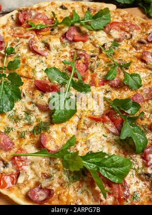 Gebackene runde Pizza mit Räucherwürsten, Pilzen, Tomaten, Käse und Laub, Essen wird in Portionen geschnitten Stockfoto