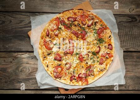 Gebackene runde Pizza mit Räucherwürsten, Pilzen, Tomaten, Käse, Speisen werden in Portionen geschnitten, Holztisch Stockfoto