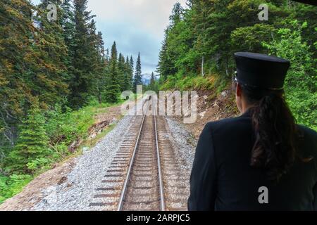 Damenleiter schaut aus dem Zug. Skagway, Alaska, Arbeiter auf der Eisenbahn im Wald. Sie trägt schwarze Uniform und professionellen Hut. Stockfoto