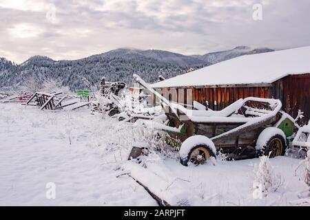 Ein alter, schneebedeckter Spreuverteiler und andere Anbaugeräte auf der Seite einer alten Holzscheune, auf einer abgelegenen Pferdefarm in Beavertail, Montana. Beavertail ist Stockfoto