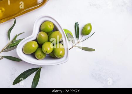 Herzförmige Schüssel voller grüner Oliven auf einem weißen Tisch, Stockfoto