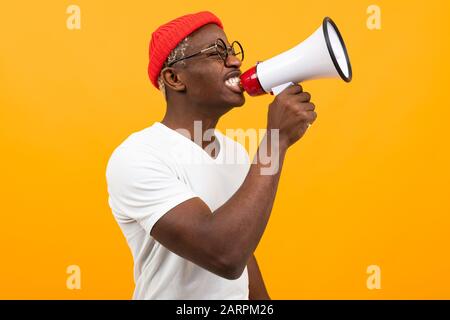 Stylischer schwarzer ansehnlicher amerikanischer Mann in weißem T-Shirt spricht Nachrichten durch ein Megafon auf isoliertem orangefarbenem Hintergrund mit Kopierraum. Stockfoto