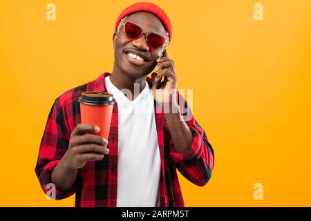 Gutaussehender schwarzer amerikaner, der auf dem Telefon spricht, während er ein Kaffeeglas zum Mitnehmen auf gelbem Studiohintergrund hält. Stockfoto