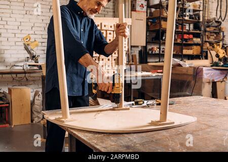 Hochrangiger männlicher Zimmermann bohrte ein Loch in Holztisch, der auf der Werkbank stand Stockfoto