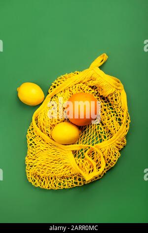 Fruchtlemons und Orange in gelber wiederverwendbarer Sack aus Mesh-Material auf grünem Hintergrund, umweltfreundliches Konzept ohne Abfall. Stockfoto