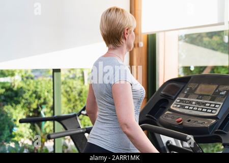 Frau mittleren Alters, die auf dem Laufband im Fitnessstudio trainiert. Gesundheitsclub mit Blick auf die Natur an sonnigen Tagen auf grünen Bäumen. Farbbild mit Kopierbereich. Stockfoto