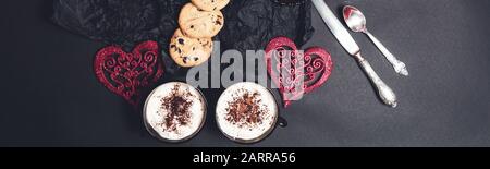 Banner mit Romantischem Frühstück. Zwei Kaffeetups, Cappuccino mit Schokoladeplätzchen und Keksen in der Nähe roter Herzen auf schwarzem Tischhintergrund. Valentine Stockfoto