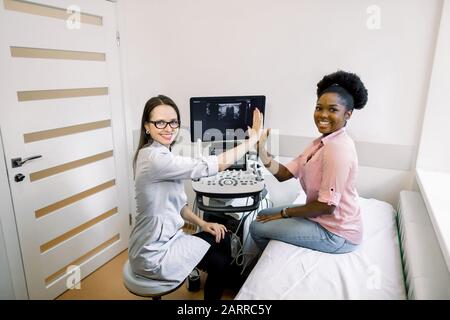 Gute Nachrichten nach der Ultraschalldiagnose. Junge afrikanische Patientin fühlt sich glücklich und lächelnd an, während sie fünf ihrer lächelnden Frau Doktor gibt, sieht Stockfoto