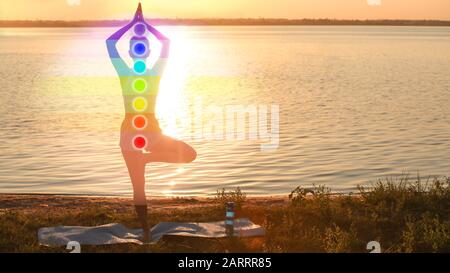 Silhouette der Frau mit gezeichneten Chakren, die Yoga in der Nähe des Flusses bei Sonnenuntergang praktizieren Stockfoto