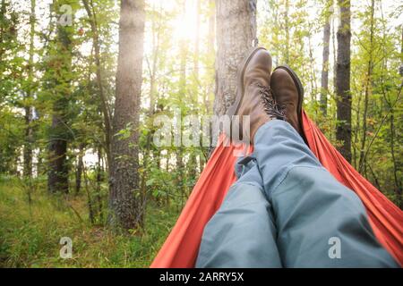 Person, die Trekkinghosen und Wanderschuhe trägt entspannende Verlegung in der Camping-Hängematte mit Blick auf den Sommerwald bei Sonnenuntergang Stockfoto