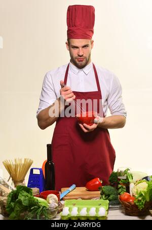 Koch in weinroten Uniformen sticht Zutaten mit Schneebesen. Küchenzubehör und Backkonzept. Mann mit Bart mit Schneebesen und Schüssel auf weißem Hintergrund. Baker steht mit geschirr und Gemüse am Tisch. Stockfoto