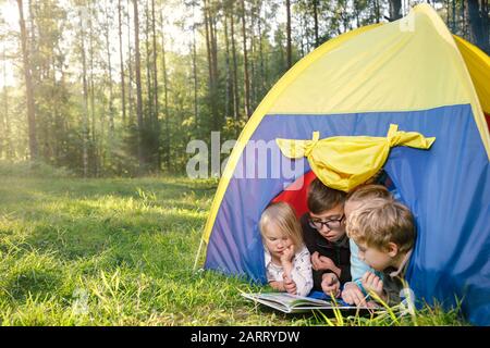 Vier Kinder unterschiedlichen Alters lesen in den Sommerferien ein Buch zusammen, das im Zelt im Sommerwald liegt Stockfoto