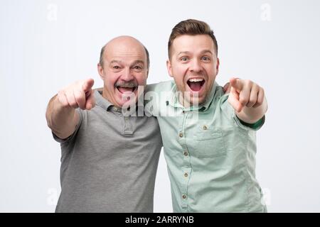 Zwei glückliche kaukasische Männer Freunde oder Vater und Sohn umarmen sich, zeigen den Finger und schauen auf die Kamera. Studio gedreht Stockfoto