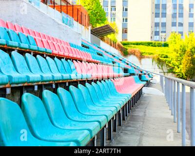 Die rhythmischen Reihen leerer türkis-roter Kunststoffstühle eines alten Sportstadions vor dem Hintergrund des Sonnenlichts im Hintergrund. Stockfoto