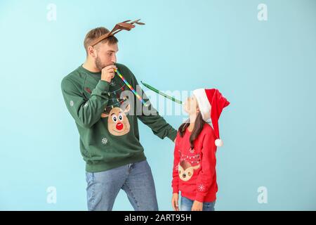 Lustige kleine Mädchen und ihr Vater in Weihnachtspullover und mit Partypfeifen auf Farbhintergrund Stockfoto