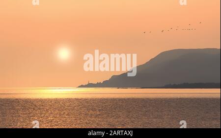 Goldener Sonnenaufgang über der Kirche am Meer zu Fuß der Berge mit Gänsescharen am Himmel Stockfoto