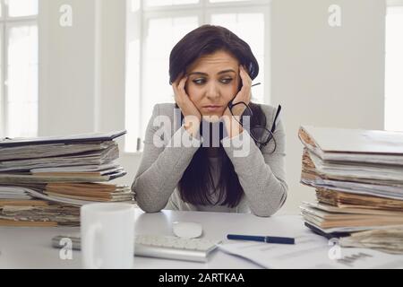 Frau brünette Geschäftsfrau in Brille mit einem Berg von Dokumenten auf dem Tisch müde depressiv empfindet enttäuscht depressiv am Tisch in Stockfoto