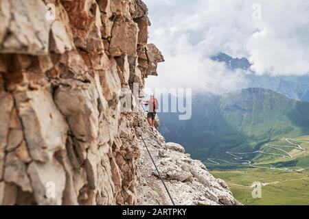 Mann auf der Via ferrata Cesare Gazzetta, Berge der Dolden, Italien, in der Nähe der Felswand, mit einer kurvenreichen Straße unten, an einem Sommertag mit Wolken versammelt Stockfoto