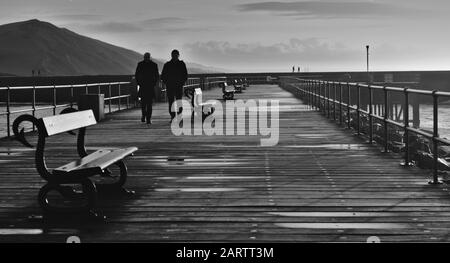 Schwarz-weiß (monochrom) Zwei Herren, die auf dem Holzsteg am Hafen von Aberystwyth mit dem Hügel Alt Wen im Hintergrund und dem spazieren gehen Stockfoto