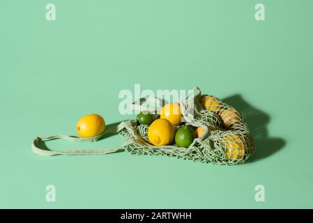 Zitronen und Limonen in einer wiederverwendbaren Einkaufstasche aus Stoff auf grünem Hintergrund. Umweltfreundliche Netztasche mit Zitrusfrüchten. Sommerfrüchte. Lebensmittel-Shopping-Konzept Stockfoto