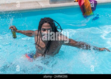 Hübsche schwarze Männer mit Dreadlocks, die eine Bierflasche halten, haben Spaß beim Spritzen von Wasser im Pool. Freizeit- und Spaßkonzept. Stockfoto