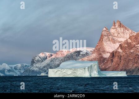 Sonnenaufgang im Fjord Scoresby Sund. Schwimmender Eisberg mit rötlich gefärbten Bergen. Kangertitittivaq, Grönland, Dänemark Stockfoto