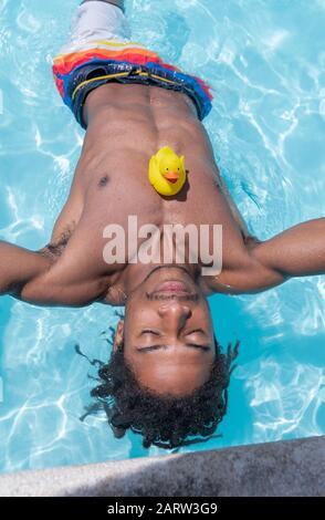 Gutaussehender schwarzer Mann mit geschlossenen Augen und einer gelben Gummiente, die auf der Brust sitzt, entspannt sich im Pool. Freizeitkonzept. Stockfoto