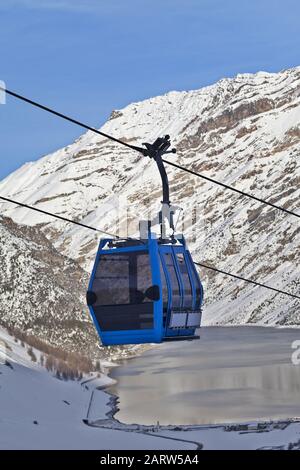 Blaue Gondelbahn, verschneite Abfahrtskiste in hohen Winterbergen, Dorf und großer See im Hintergrund. Italienische Alpen am Abend. Livigno, Region von Stockfoto