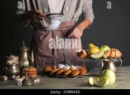 Frau streut Zuckerpulver auf leckere Plätzchen auf den Tisch Stockfoto
