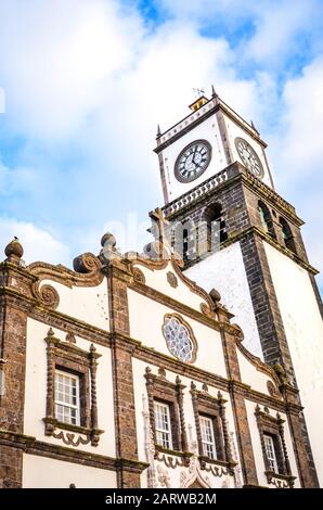 Die Außenfassade der St. Sebastian Kirche, Igreja Matriz de Sao Sebastiao, in Ponta Delgada, Azoren, Portugal. Weißer Uhrturm von unten. Blauer Himmel und Wolken darüber. Sonnenuntergang, vertikales Foto. Stockfoto