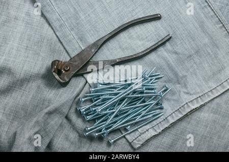 Eine Handvoll metallischer selbstschneiderschrauben und sehr gealterte und rostige Zangen liegen auf dem Hintergrund eines alten Denim-Stoffes. Makro mit selektivem Fokus aufgenommen mit Stockfoto
