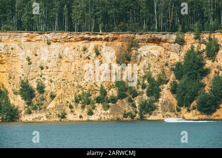 Hohe felsige sandige Steilufer mit Pinien bedeckt über einem klaren und blauen See. Schnelle Motorboot schneidet durch die Wasseroberfläche Stockfoto