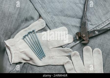 Eine Handvoll metallischer Selbstschneiderschrauben und sehr gealterte und rostige Zangen liegen auf Safety Strickhandschuhen für den industriellen Einsatz vor dem Hintergrund Stockfoto