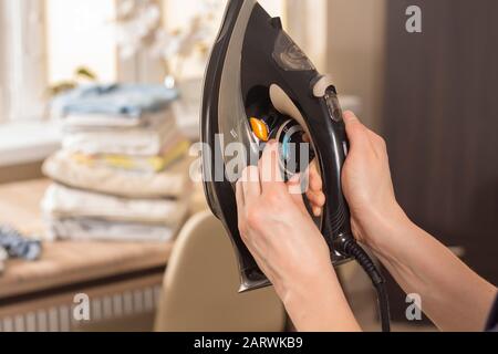 Die Hand einer Frau hält das Bügeleisen und stellt die richtige Bügeltemperatur ein. Gebügelte, gefaltete Knöchelbekleidung liegt im Hintergrund auf dem Tisch. Stockfoto