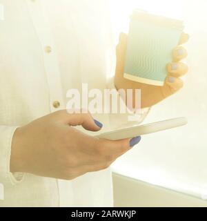 Ein Mädchen mit blauer Maniküre hält ein Smartphone und eine Papierbecher Kaffee im Sonnenlicht in den Händen. Stockfoto