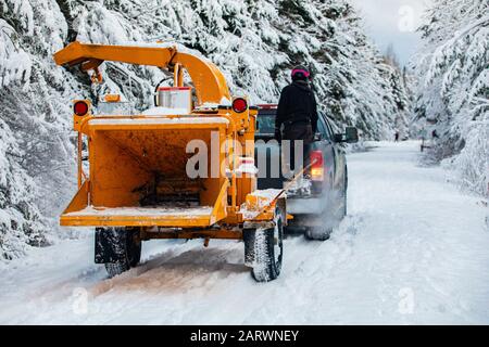 Ein weiter Blick auf einen Arboriculturisten, Baumchirurgen oder Landschaftsgärtner, der einen mobilen Holzschipper mit einem Pickup-Truck auf einer schneebedeckten Straße hinunterschleppt Stockfoto