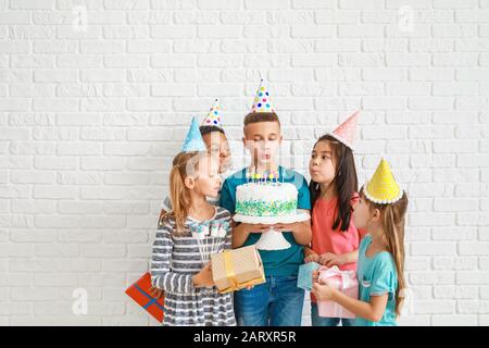 Kleine Kinder bläsen Kerzen auf Geburtstagskuchen auf weißem Backstein-Hintergrund aus Stockfoto