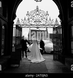 Ein Bride und Ein Bräutigam, die die Kirche verlassen, als sie mit ihrem Chauffeur und dem Auto heiraten, das auf sie draußen wartet Stockfoto